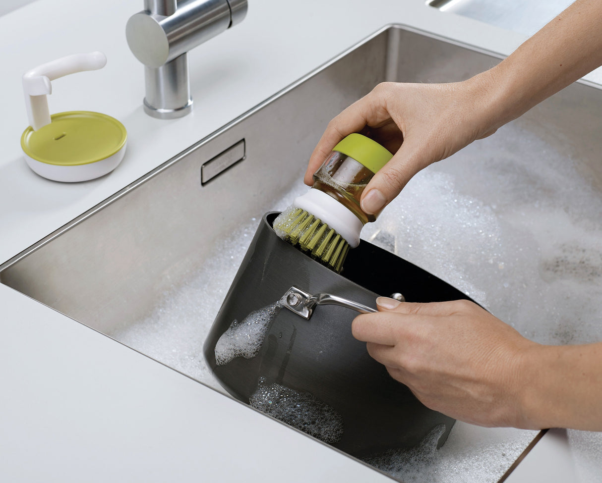 Thumb pressing the top button to release washing-up liquid through the bristles.