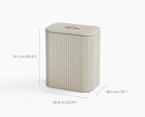 The ecru Tota Luxe laundry basket styled beside a bed in a bedroom setting.