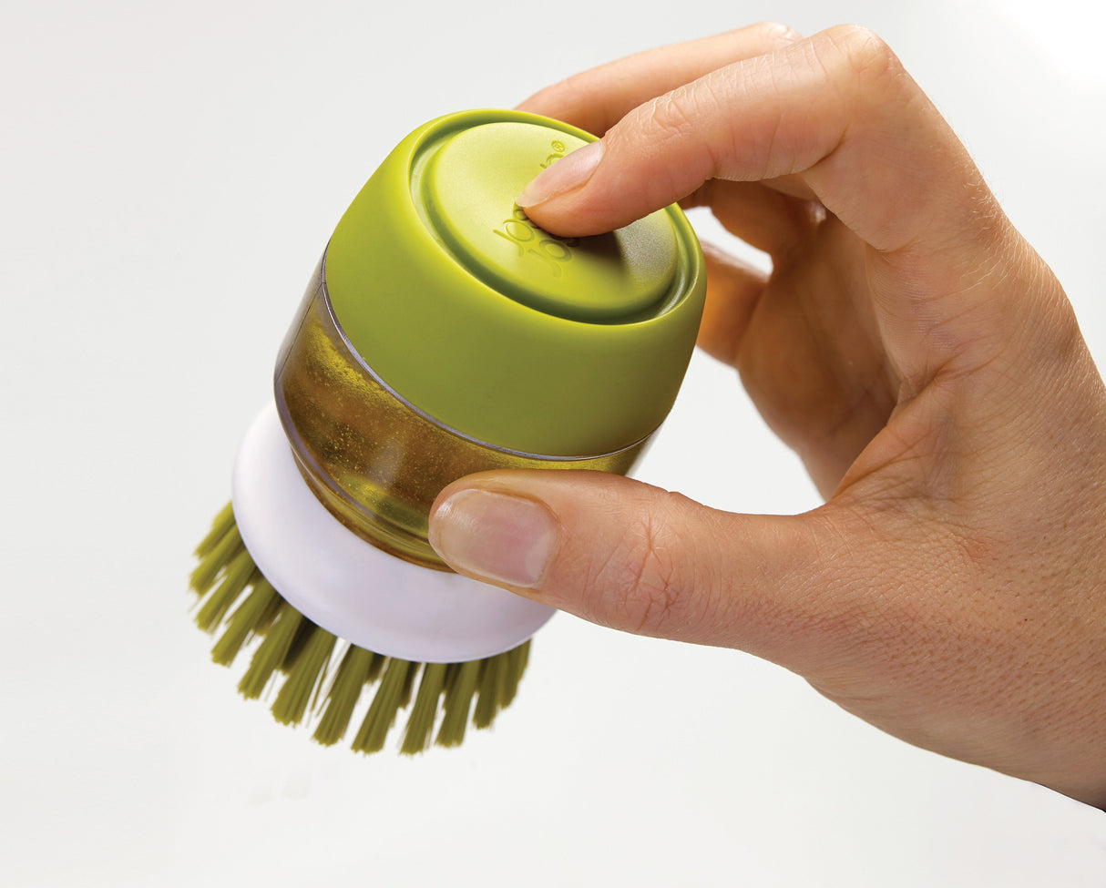The palm-sized green brush held in the hand while scrubbing a pan.